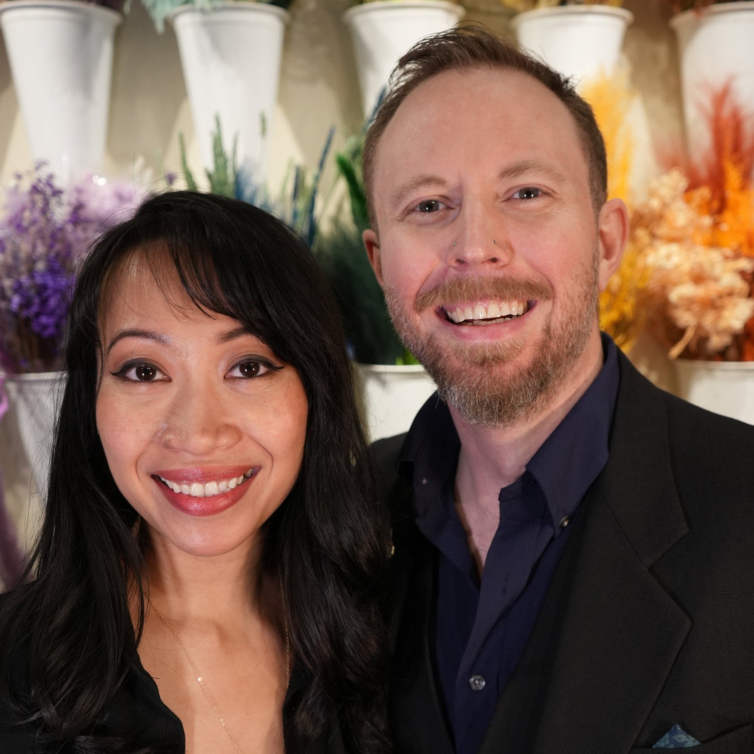 Two people smiling in front of decorative vases with floral arrangements.